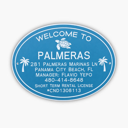 A blue oval sign with white text reads: Welcome to Palmeras. Below are an address, contact details, and a license number: 281 Palmeras Marinas Ln, Panama City Beach, FL, Manager: Flavio Yepo, 480-414-8648, Short Term Rental License #CND1308113.