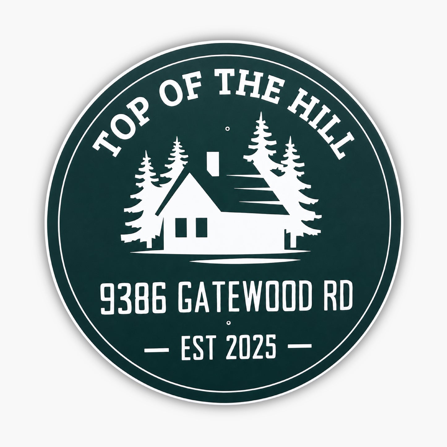 Round green sign with white design and text: TOP OF THE HILL at the top, featuring a house silhouette with trees. Below, text reads 9386 GATEWOOD RD and EST 2025. Set against a light-colored siding background.