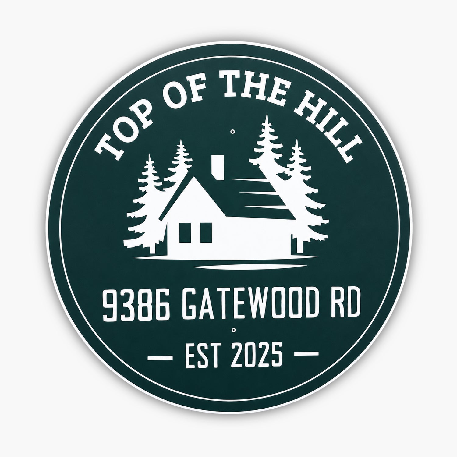 Round green sign with white design and text: TOP OF THE HILL at the top, featuring a house silhouette with trees. Below, text reads 9386 GATEWOOD RD and EST 2025. Set against a light-colored siding background.