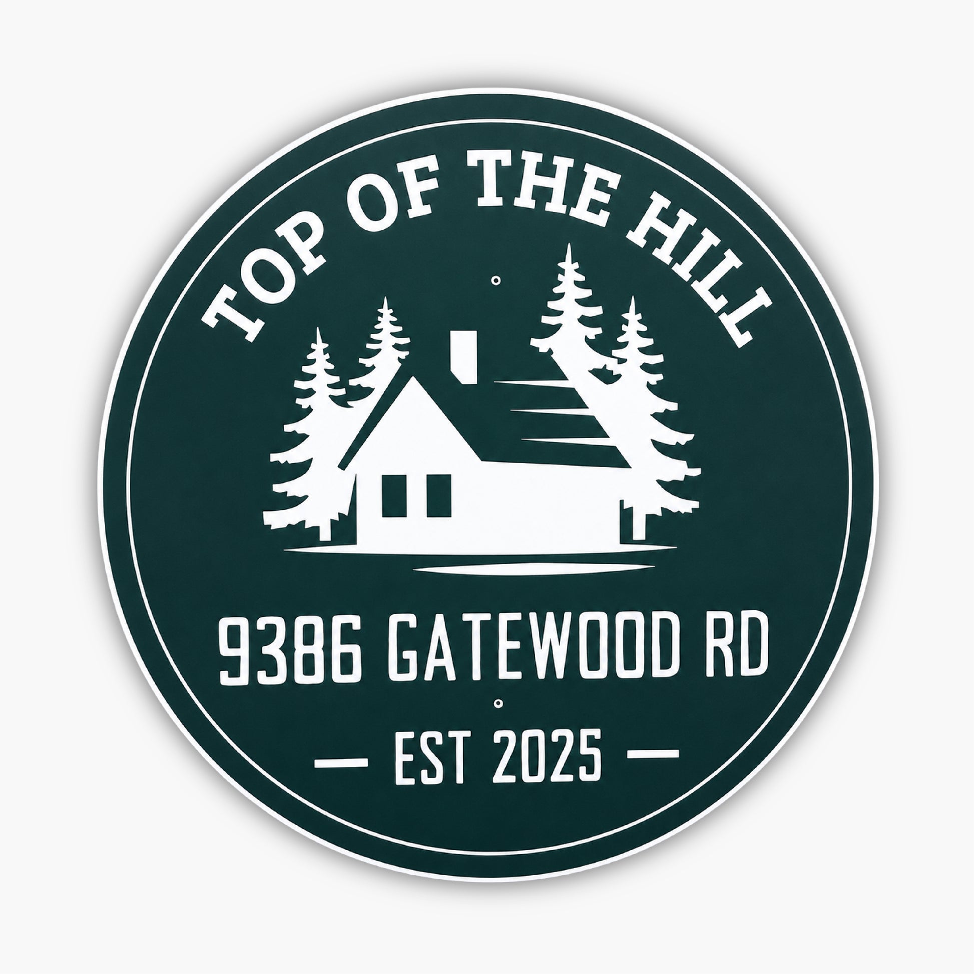 Round green sign with white design and text: TOP OF THE HILL at the top, featuring a house silhouette with trees. Below, text reads 9386 GATEWOOD RD and EST 2025. Set against a light-colored siding background.