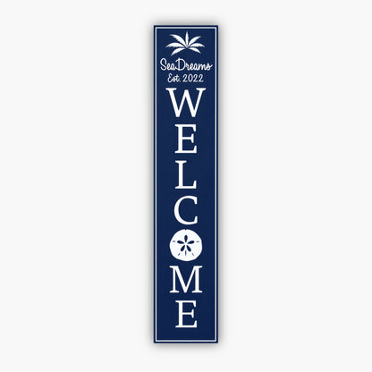 Tall blue vertical welcome sign with white text reading Sea Dream and Est. 2022 at the top, with a palm tree graphic. Large vertical WELCOME text follows, featuring a sand dollar graphic within the O. The background is a beige siding wall.