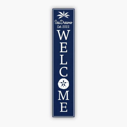 Tall blue vertical welcome sign with white text reading Sea Dream and Est. 2022 at the top, with a palm tree graphic. Large vertical WELCOME text follows, featuring a sand dollar graphic within the O. The background is a beige siding wall.