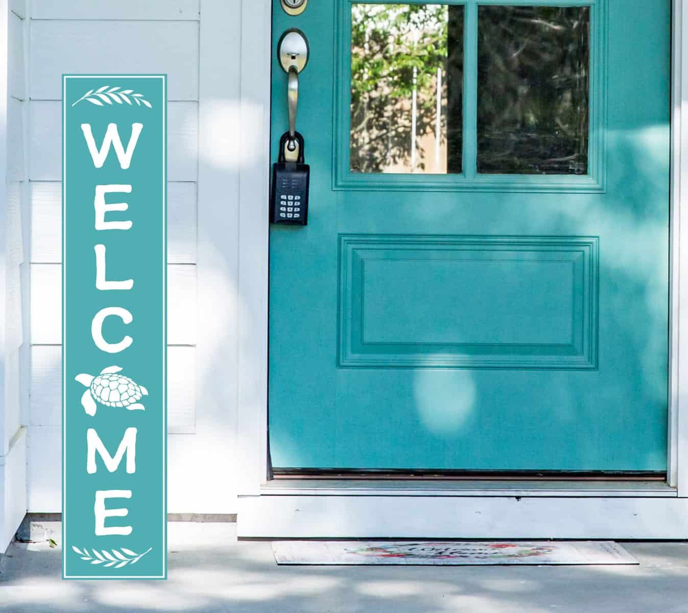A teal front door with a keypad lock is flanked by a Personalized Vertical Beach House Welcome Sign featuring a turtle design, casting dappled shadows on the white siding and porch as sunlight filters through leaves.