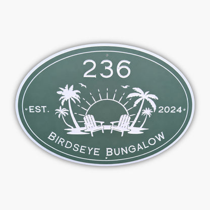 The Personalized Oval Custom Beach House Sign is green with white text, displaying 236 Birdseye Bungalow and Est. 2024. It features a sun setting behind two palm trees and lounge chairs, surrounded by birds.