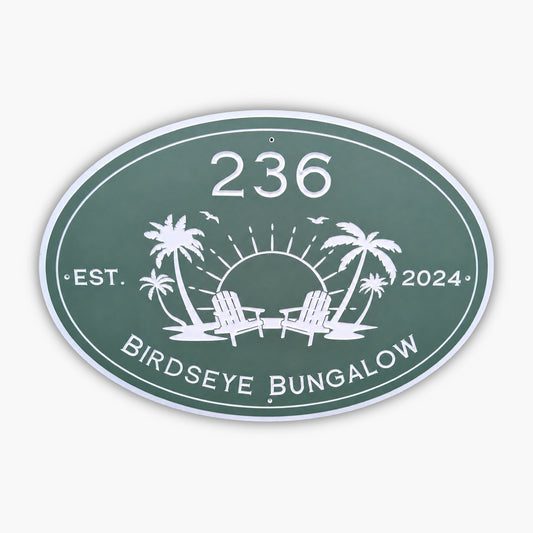The Personalized Oval Custom Beach House Sign is green with white text, displaying 236 Birdseye Bungalow and Est. 2024. It features a sun setting behind two palm trees and lounge chairs, surrounded by birds.