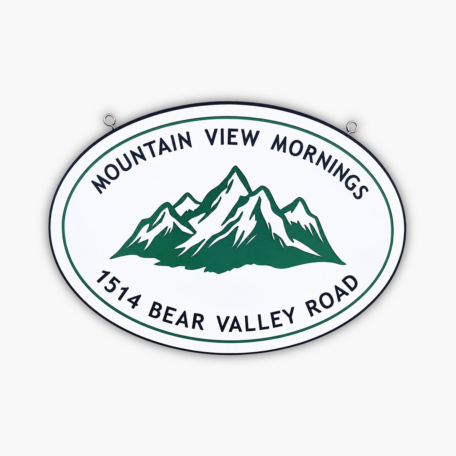 An oval sign with a green mountain illustration and text that reads MOUNTAIN VIEW MORNINGS above and 1514 BEAR VALLEY ROAD below, mounted on a beige wall with two hooks at the top.