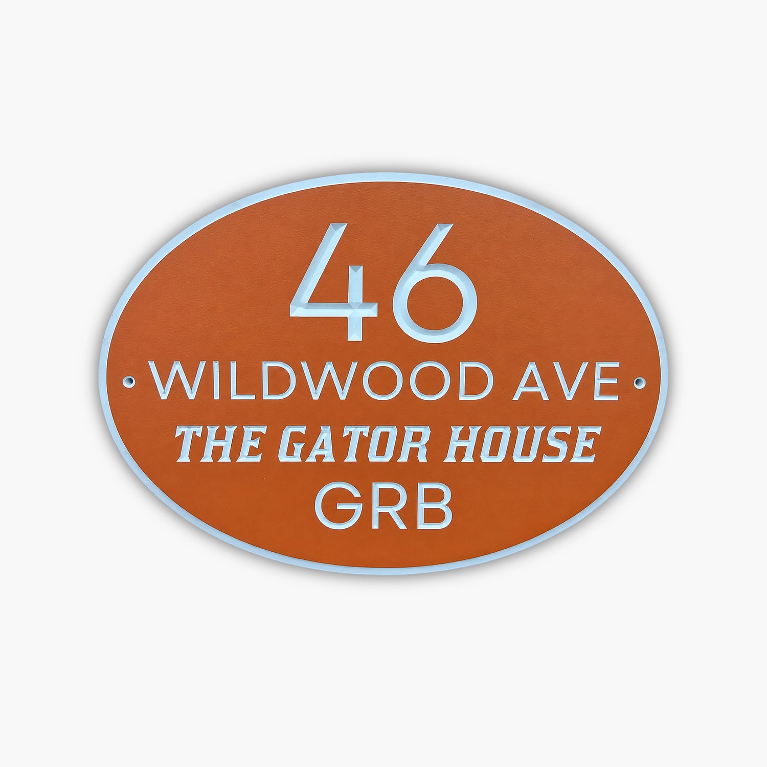 The Personalized Address Sign features an oval design with an orange background and white border. It reads 46 Wildwood Ave, The Gator House, GRB in white text, evoking the charm of a cozy cabin sign against a gray wooden wall.