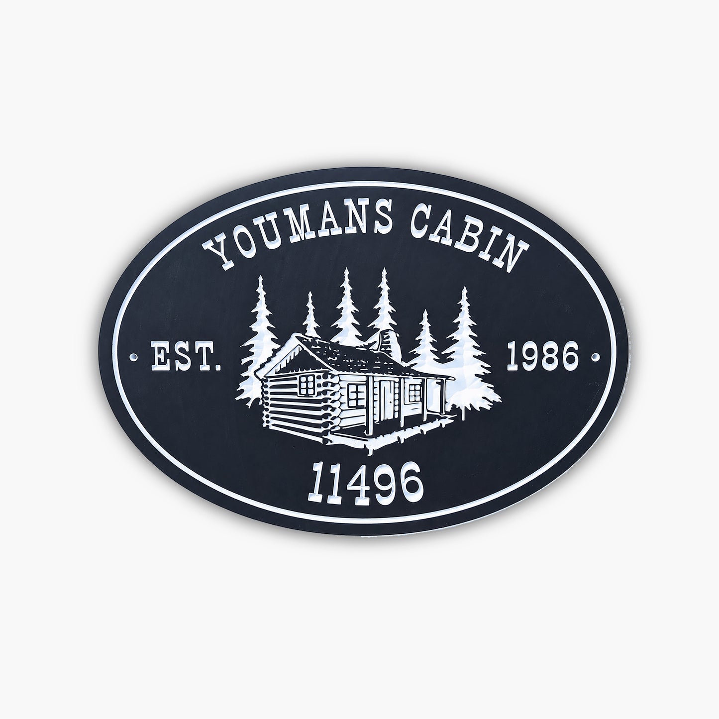 An oval sign reads YOUMANS CABIN EST. 1986 11496 with an illustration of a log cabin surrounded by trees, all in white on a dark background. The sign rests against light-colored siding.