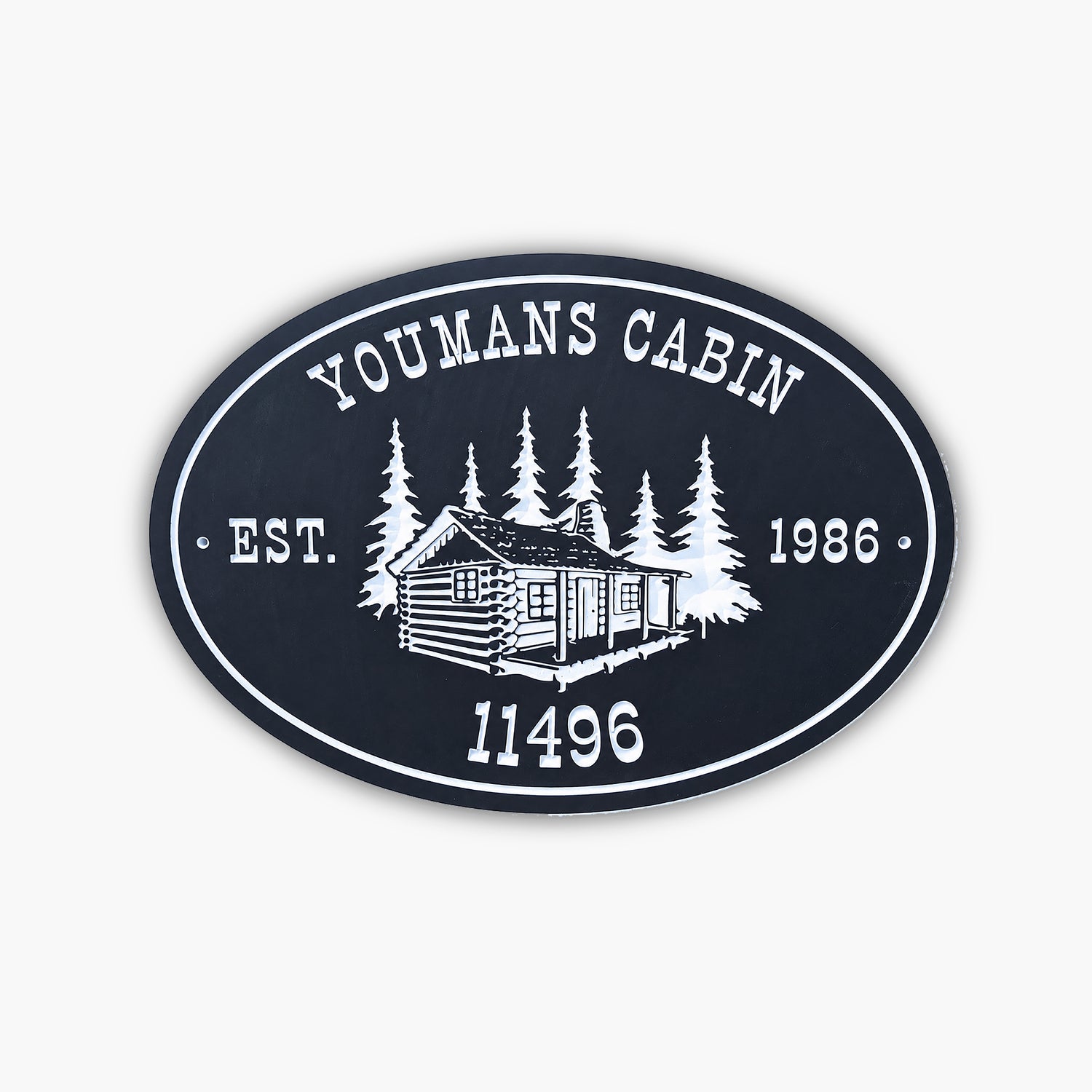 An oval sign reads YOUMANS CABIN EST. 1986 11496 with an illustration of a log cabin surrounded by trees, all in white on a dark background. The sign rests against light-colored siding.