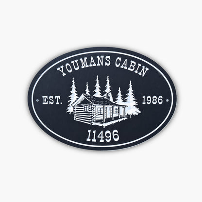 An oval sign reads YOUMANS CABIN EST. 1986 11496 with an illustration of a log cabin surrounded by trees, all in white on a dark background. The sign rests against light-colored siding.