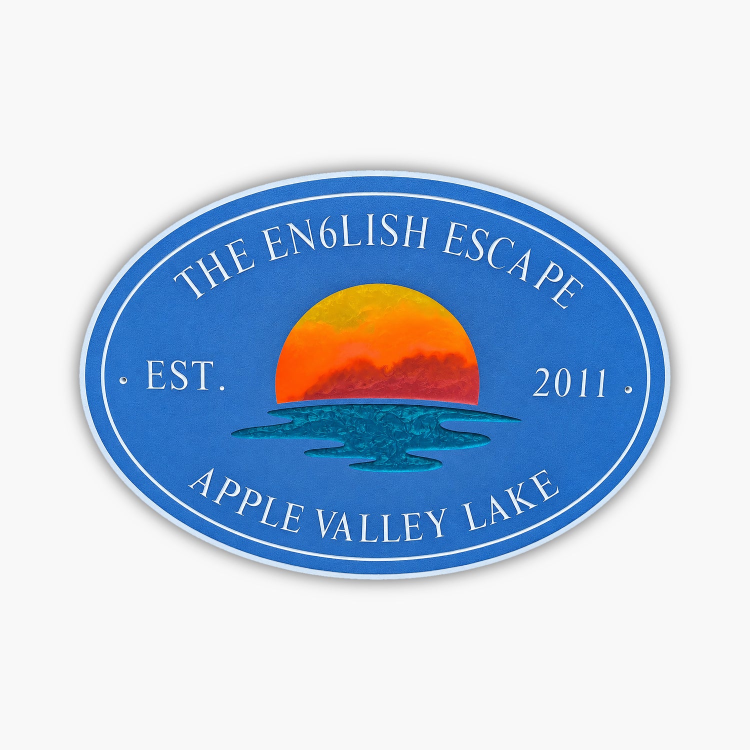 Blue oval sign with white text: The English Escape, Est. 2011, Apple Valley Lake. Like a gallery of natures artistry, it features a vivid orange and yellow sunset over a stylized body of water in the center.
