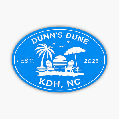 The Personalized Oval Beach House Sign is blue and white, featuring a tropical scene with a palm tree, two beach chairs, an umbrella, and a sunset over water. It reads Dunns Dune, Est. 2023, KDH, NC, making it ideal for your vacation rental sign!.