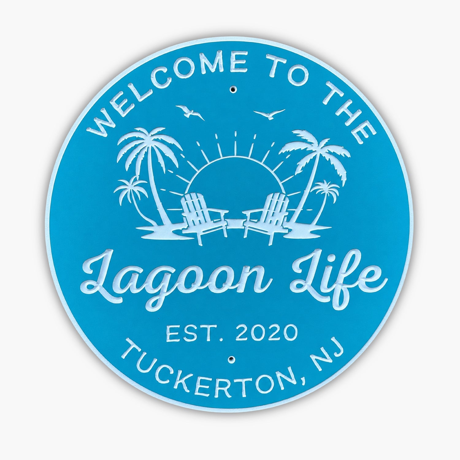 Round blue sign with white text and graphics, resembling an art gallery piece. It reads Welcome to the Lagoon Life, featuring palm trees, Adirondack chairs, a sun with rays, birds, and Est. 2020 Tuckerton, NJ.
