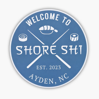 A round blue Personalized Custom Round Beach House Sign features the white text Welcome to Shore Shi, embellished with sushi rolls, crossed chopsticks, Est. 2023, and Ayden, NC.