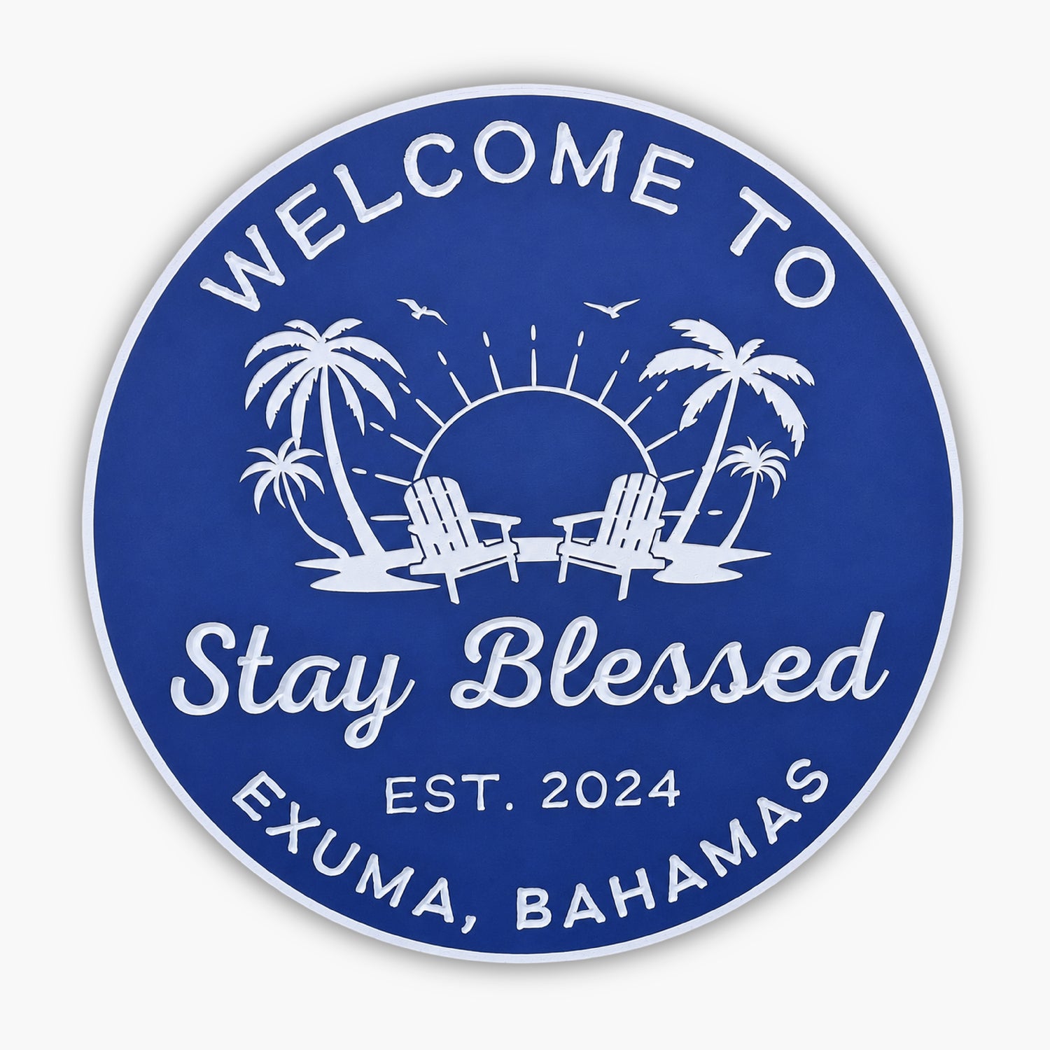A round blue sign with white text reads Welcome to Stay Blessed Gallery, Est. 2024, Exuma, Bahamas. It showcases two palm trees, two beach chairs, a setting sun, and birds in flight.
