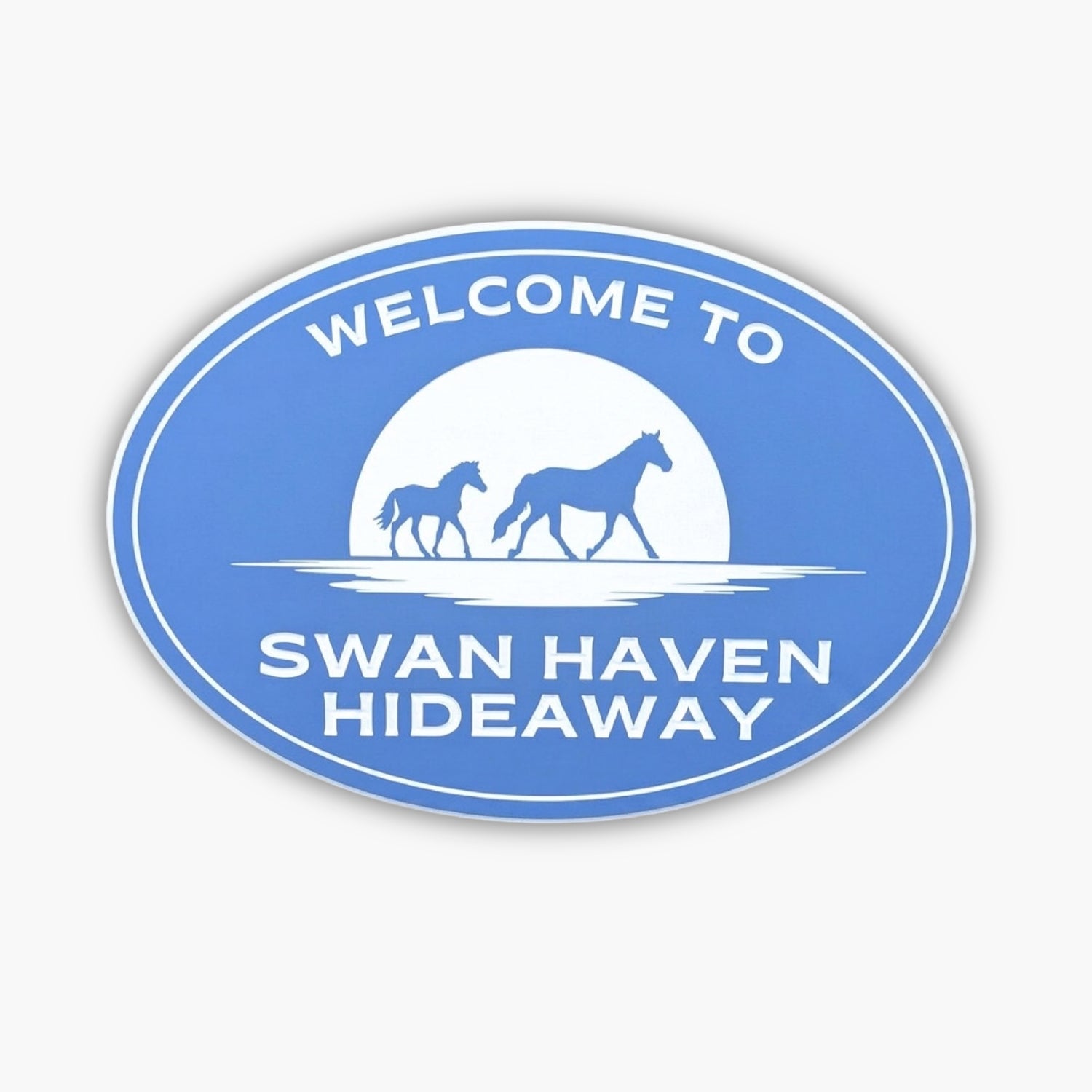 Blue oval sign featuring silhouettes of two horses walking in front of a sun. The text, Welcome to Swan Haven Hideaway, is elegantly displayed in white capital letters, inviting you to explore the tranquil gallery nestled within this serene retreat.