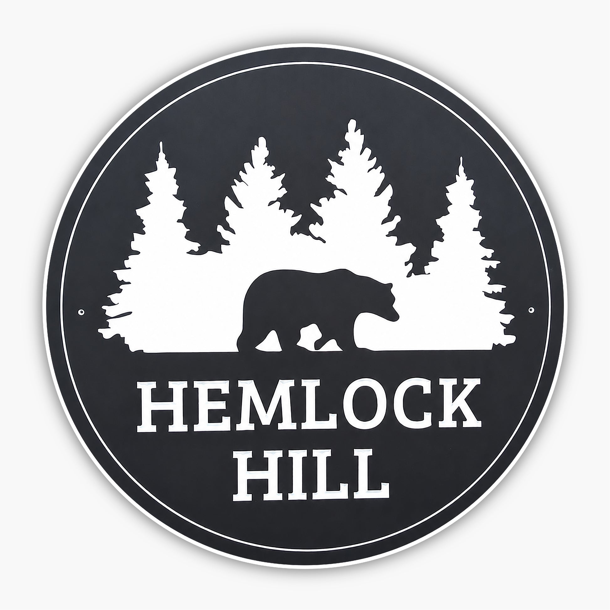 A circular black sign with white silhouettes of pine trees and a bear. The text HEMLOCK HILL is written in bold white letters below the bear and trees. The sign is mounted on a wooden surface with siding in the background.
