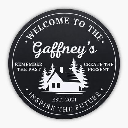 The Personalized Round Cabin Sign features a black circular background with white text: Welcome to the Gaffneys at the top. A custom cabin silhouette and trees grace the center, flanked by Remember the Past, Create the Present. Below is Est. 2021, with Inspire the Future at the bottom.
