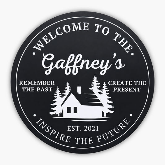 The Personalized Round Cabin Sign features a black circular background with white text: Welcome to the Gaffneys at the top. A custom cabin silhouette and trees grace the center, flanked by Remember the Past, Create the Present. Below is Est. 2021, with Inspire the Future at the bottom.