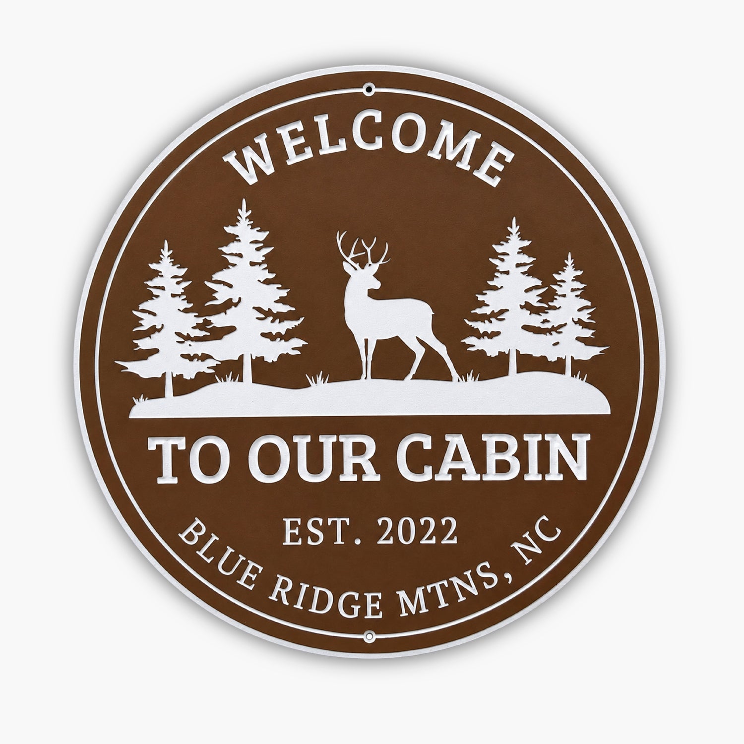 Round brown sign with white text and graphics, resembling an art gallery piece. It says, Welcome To Our Cabin Est. 2022 Blue Ridge Mtns, NC, and features silhouettes of three pine trees and a deer.
