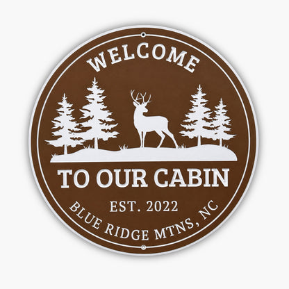 Round brown sign with white text and graphics, resembling an art gallery piece. It says, Welcome To Our Cabin Est. 2022 Blue Ridge Mtns, NC, and features silhouettes of three pine trees and a deer.