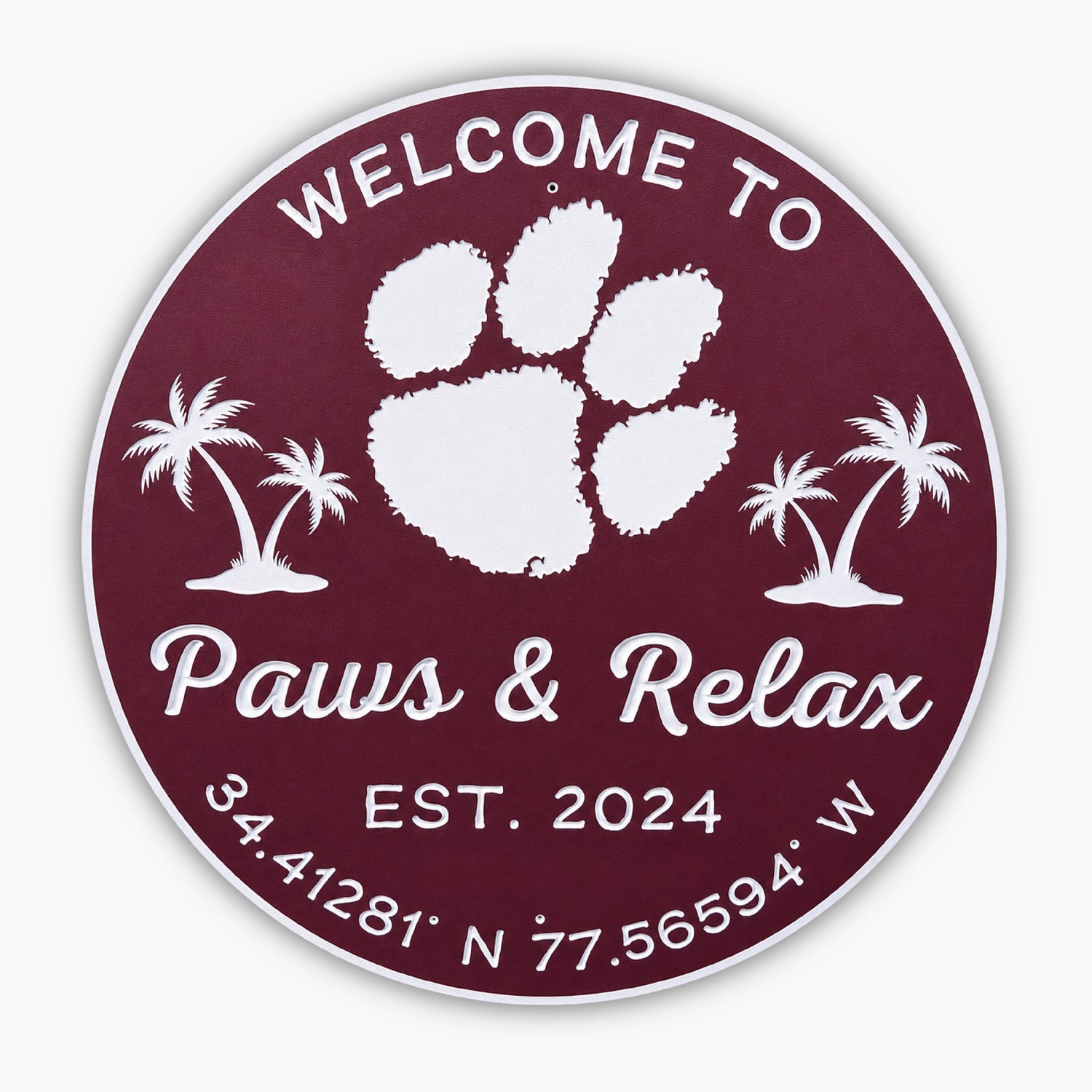 A circular sign with a burgundy background showcases a large white paw print and palm trees, reminiscent of an art gallerys emblem. Text reads: Welcome to Paws & Relax, Est. 2024. Coordinates are 34.41281° N, 77.56594° W.
