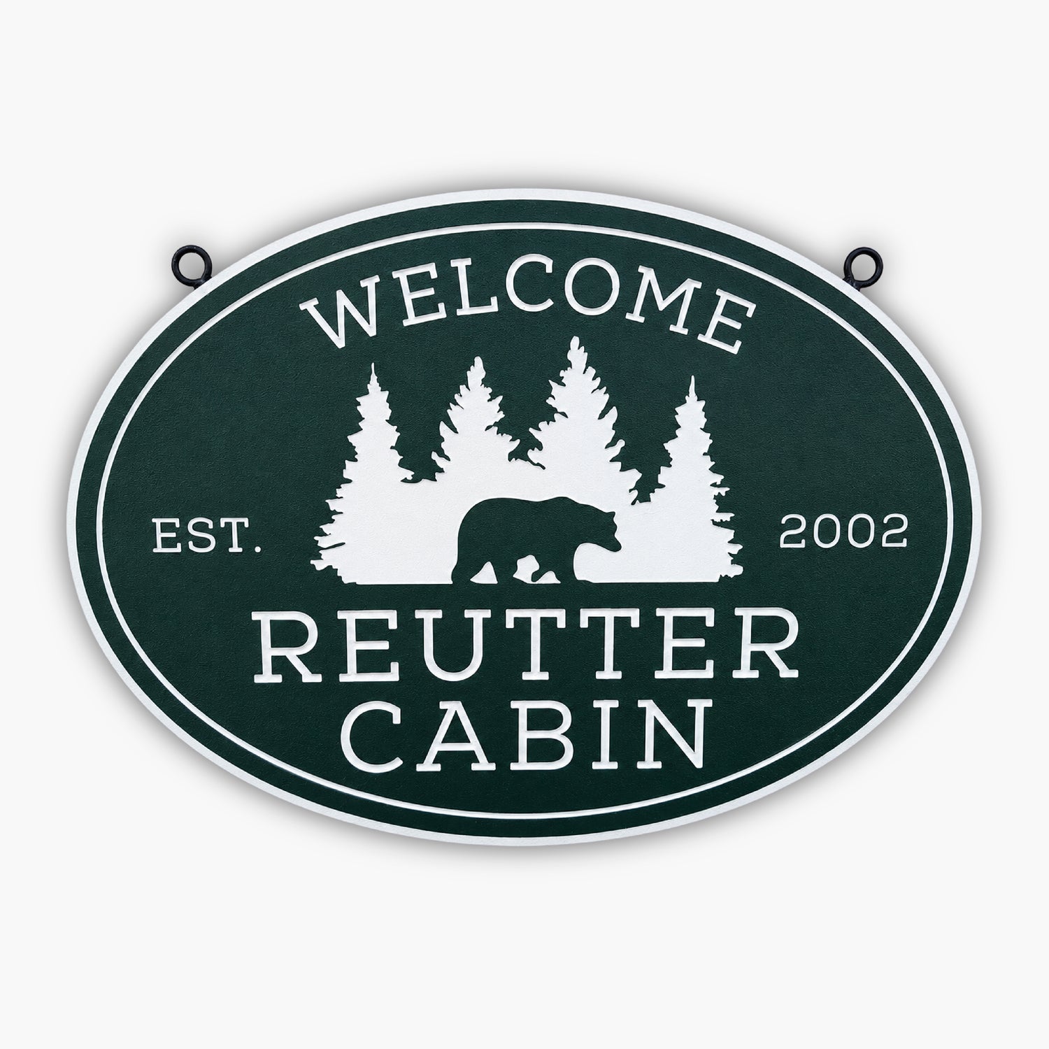 Oval sign with a green background and white text reads Welcome at the top. Below, a silhouette of a bear walking in front of pine trees. Like artwork fit for a gallery, the text underneath says Reutter Cabin with Est. 2002 on each side.