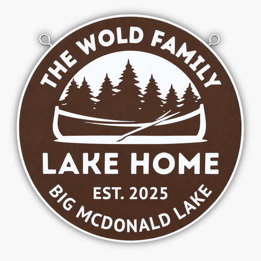 A round brown sign with white text reads The Wold Family Lake Home, Est. 2025, Big McDonald Lake. The sign features a canoe and pine trees in the center.