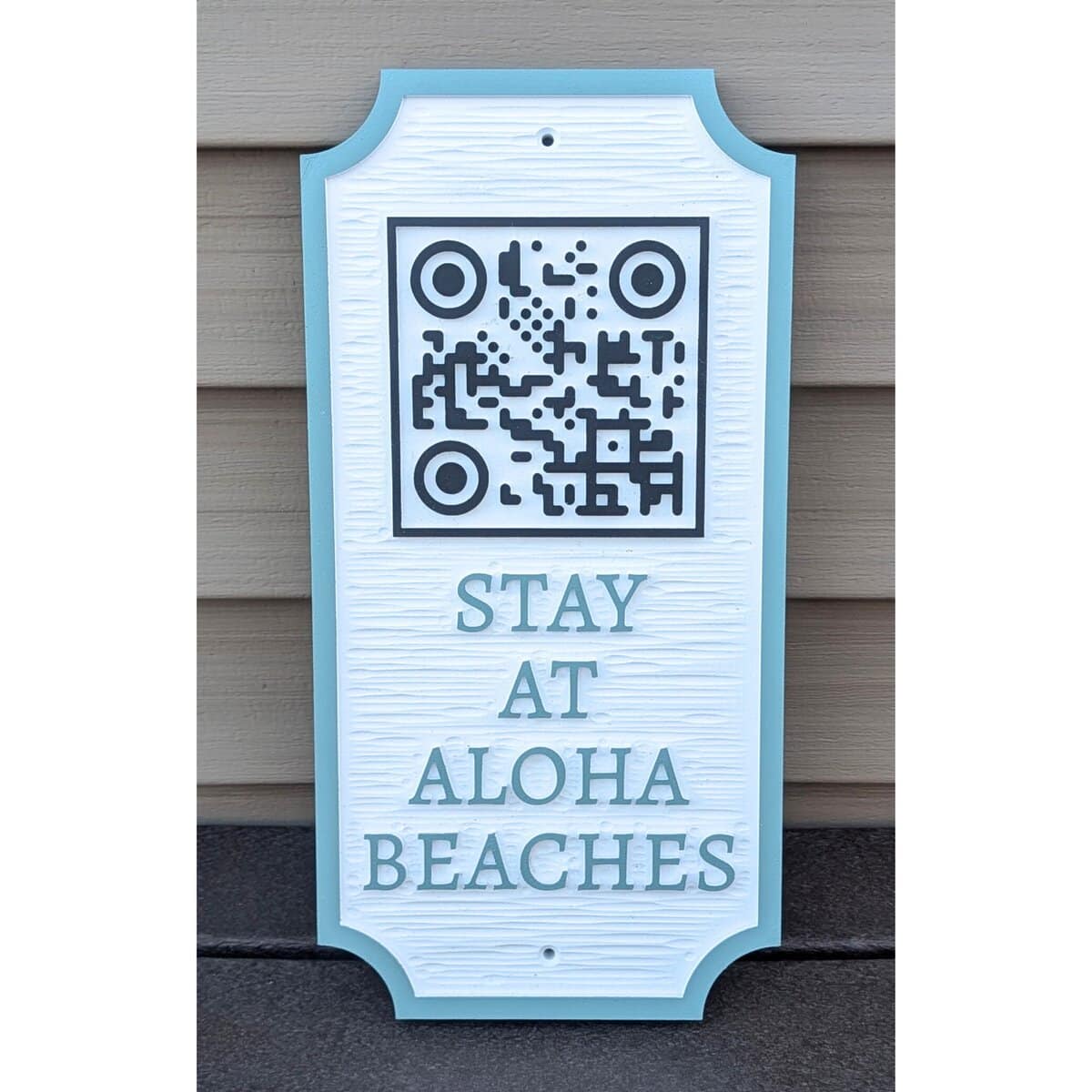 A decorative sign with a QR code at the top and the words STAY AT ALOHA BEACHES below, leaning against a beige paneled wall.