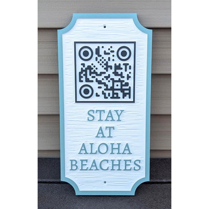 A decorative sign with a QR code at the top and the words STAY AT ALOHA BEACHES below, leaning against a beige paneled wall.